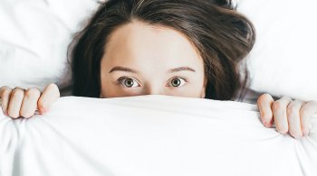 Woman peeking from under white covers, wide awake—symbolizing the struggle to get better sleep naturally