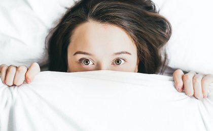 Woman peeking from under white covers, wide awake—symbolizing the struggle to get better sleep naturally