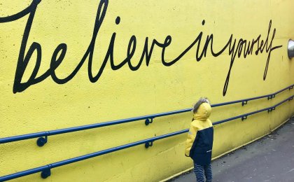 Child in yellow coat reading mural that says ‘Believe in yourself,’ symbolizing mindset shift and inner strength
