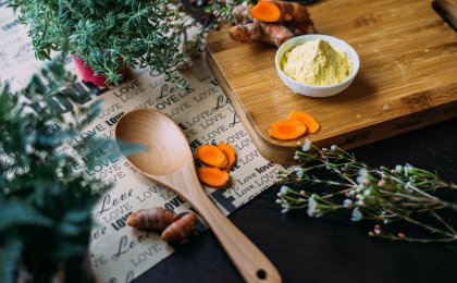 Fresh turmeric root and powder on a wooden cutting board surrounded by herbs and a spoon