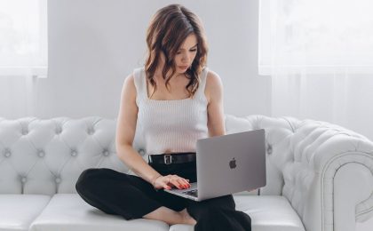 Woman sitting on a couch with a laptop open on her lap, reading in a relaxed home setting.